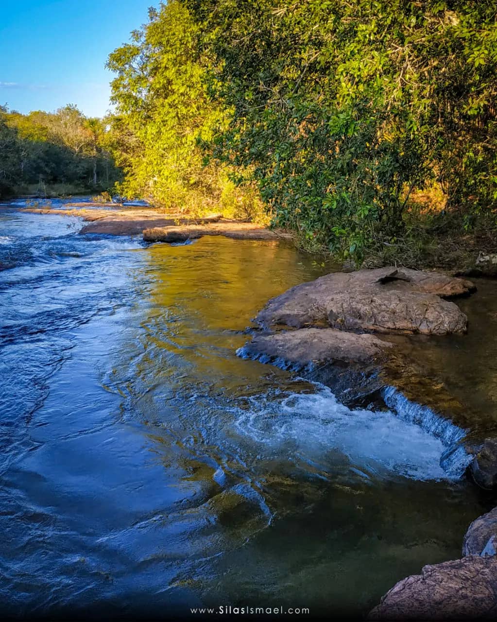 Rio correndo entre pedras, com mata iluminada pelo sol da tarde